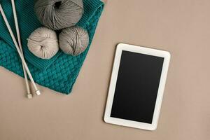 gris y turquesa tejido de punto lana, tejido de punto agujas y un tableta con un negro pantalla en beige antecedentes. parte superior vista. Copiar espacio foto