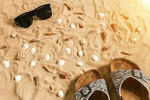 Black flip-flops and sunglass, seashell on sand. With place for your text. Top view photo