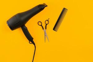 Black hair dryer, comb and scissors on yellow paper background. Top view photo