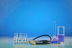 Beaker, medical flask. Purple chemical reagent. Six ampoules with liquid, syringe and phonendoscope, blue background. Worldwide Coronavirus pandemic photo