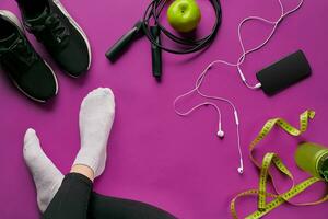 Flat lay gym equipment such as jump rope, bottle of waters, smartphone with headphone, apple and human legs in socks on color background. photo