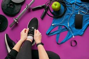 Young woman laces sneakers, preparing for training. Bottle of water, yoga mat, phone, headphones on purple background flat lay top view. photo