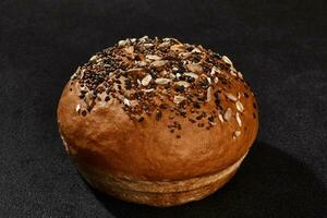 Fresh, palatable baked bun sprinkled with sesame and sunflower seeds against black background with copy space. Rural cuisine or bakery. Close-up photo