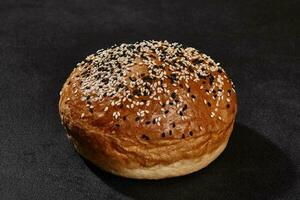 Fresh, tasty baked bun sprinkled with sesame seeds against black background with copy space. Rural cuisine or bakery. Close-up photo