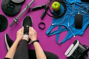 Young woman laces sneakers, preparing for training. Bottle of water, yoga mat, phone, headphones on purple background flat lay top view. photo