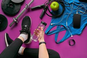 Girls legs wearing black trainers sitting on a mat surrounded by mobile phone, earphones, towel and a shaker in the fitness club. Top view. photo