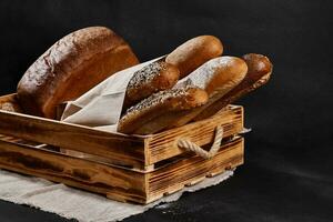 Assortment of fresh baked bread in a paper bag and wooden box standing on a burlap against black background with copy space. Close-up photo