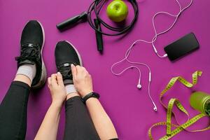 Young woman laces sneakers, preparing for training. Bottle of water, yoga mat, phone, headphones on purple background flat lay top view. photo