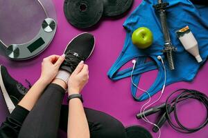 Young woman laces sneakers, preparing for training. Bottle of water, yoga mat, phone, headphones on purple background flat lay top view. photo