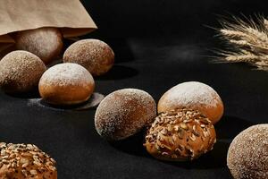 Assortment of fresh baked buns with sesame and sunflower seeds, sprinkled with flour, in a paper bag. Black background, bunch of wheat. Close-up photo