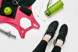 Girls legs wearing black trainers sitting on a mat surrounded by mobile phone, earphones, towel and a shaker in the fitness club. Top view. photo