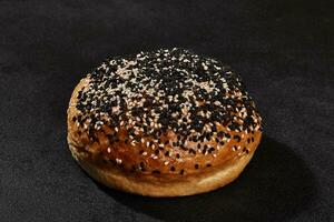 Fresh, palatable baked bun sprinkled with sesame seeds against black background with copy space. Rural cuisine or bakery. Close-up photo