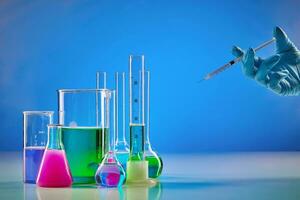 Hand in disposable glove holding syringe. Colorful reagents in beakers, medical flasks and measuring cylinder, blue background. Coronavirus pandemic photo