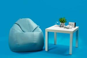 Bean bag, white coffee table with candles on it and in glass candlestick, green flower in pot, glasses, decorative iron triangle. Blue background photo