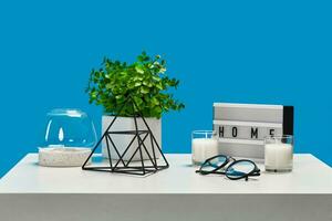 White table with candles on it and in a glass candlestick, green plant in pot, glasses, iron triangle, home decor element. Blue background. Close up photo