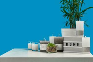 White table with green plant and succulent in pots, some different sized and colors candles, lamp with inscription home. Blue background. Close up photo
