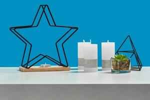 White table with green succulent, three big candles and one small in candlestick in form of iron star, black triangle. Blue background. Close up photo