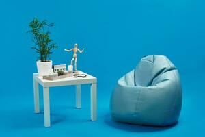 Turquoise bean bag, white coffee table with green flower in pot, candles, succulent, wooden figurine of human, glasses. Blue background. Copy space photo