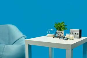 Bean bag, white coffee table with candles on it and in glass candlestick, green flower in pot, glasses, decorative iron triangle. Blue background photo