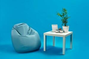 Turquoise armchair bean bag and white coffee table with green flower in a pot, three candles and photo frame on it. Blue studio background. Copy space