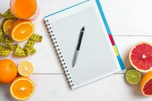 Fresh juice in glass from citrus fruits - lemon, grapefruit, orange, notebook with pencil on white wooden background photo
