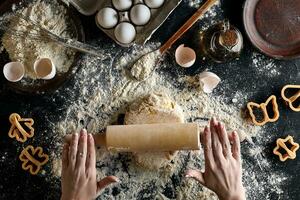 Female hands rolling dough with a rolling pin on a black table. Top view photo