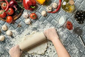 Female hands roll out dough on metal background, close up. Chef makes dough. Table with vegetables and kitchenware. Italian cuisine concept. Pizza photo