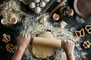 Female hands rolling dough with a rolling pin on a black table. Top view photo