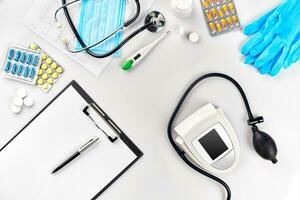 White electric tonometer with stethoscope, multicolored pills, mask and thermometer on white table. Top view photo