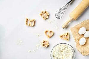 Bowl with wheat flour, rolling pin, whisk, eggs, cookie cutters. Top view on a white table with a copy space photo