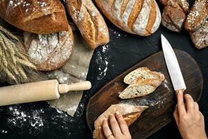 Woman hands cutting a loaf of bread on rustic wooden board, with wheat ears and knife, top view. photo