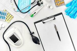 White electric tonometer with stethoscope, multicolored pills, mask and thermometer on white table. Top view photo