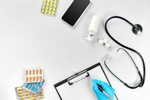 Workplace of a doctor. Pills in hand, stethoscope, clipboard and glasses on white table photo