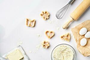 Bowl with wheat flour, rolling pin, whisk, eggs, cookie cutters. Top view on a white table with a copy space photo