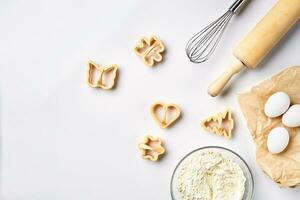 Bowl with wheat flour, rolling pin, whisk, eggs, cookie cutters. Top view on a white table with a copy space photo