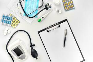 White electric tonometer with stethoscope, multicolored pills, mask and thermometer on white table. Top view photo