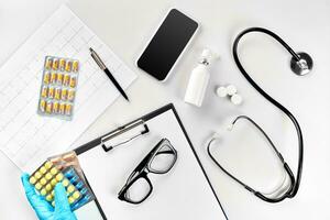 Workplace of a doctor. Pills in hand, stethoscope, clipboard and glasses on white table photo