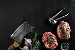 Two raw fresh ribeye steaks with salt, pepper, rosemary, garlic and an ax on black background photo