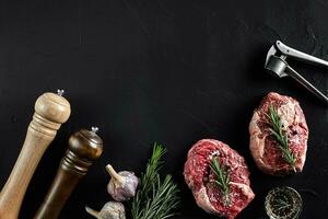 Two raw fresh ribeye steaks with salt, pepper, rosemary and garlic on black background photo