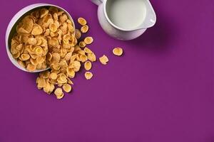 Bowl with corn flakes, jug of milk on purple background, top view photo