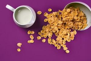 Bowl with corn flakes, jug of milk on purple background, top view photo