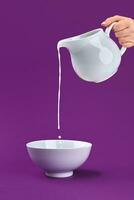 Partial view of hand pouring fresh organic milk from jug to bowl on purple background photo