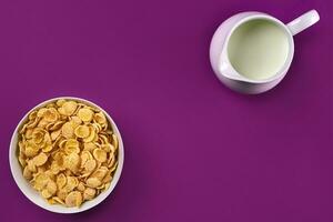 Bowl with corn flakes, jug of milk on purple background, top view photo