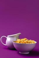 Bowl with corn flakes, jug of milk on purple background. oncept of a healthy breakfast photo