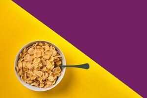 Bowl with corn flakes and spoon on purple and yellow background, top view photo