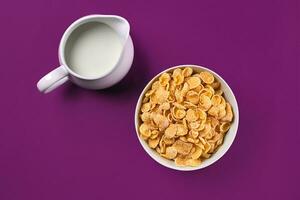 Bowl with corn flakes, jug of milk on purple background, top view photo