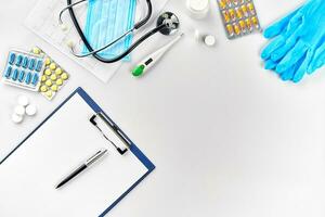 Medical equipment pills, mask, blue gloves, thermometer and stethoscope, white blank with a pen on white background. Top view. Copy space photo