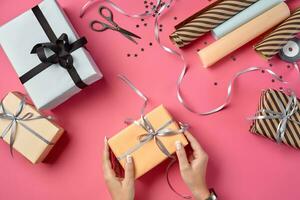 Hands of a woman are holding rose paper gift box tied with silver ribbon and bow against a pink background. Close-up, copy space, top view. photo