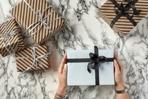 Hands of a woman are holding white paper gift box tied with black ribbon and bow against a marble background. Close-up, copy space, top view. photo