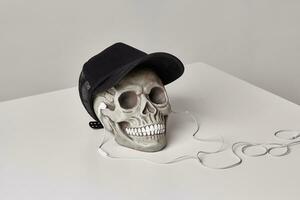 Realistic model of a human skull with teeth in a black baseball cap and headphones on a light table, white background. Halloween horror concept. photo